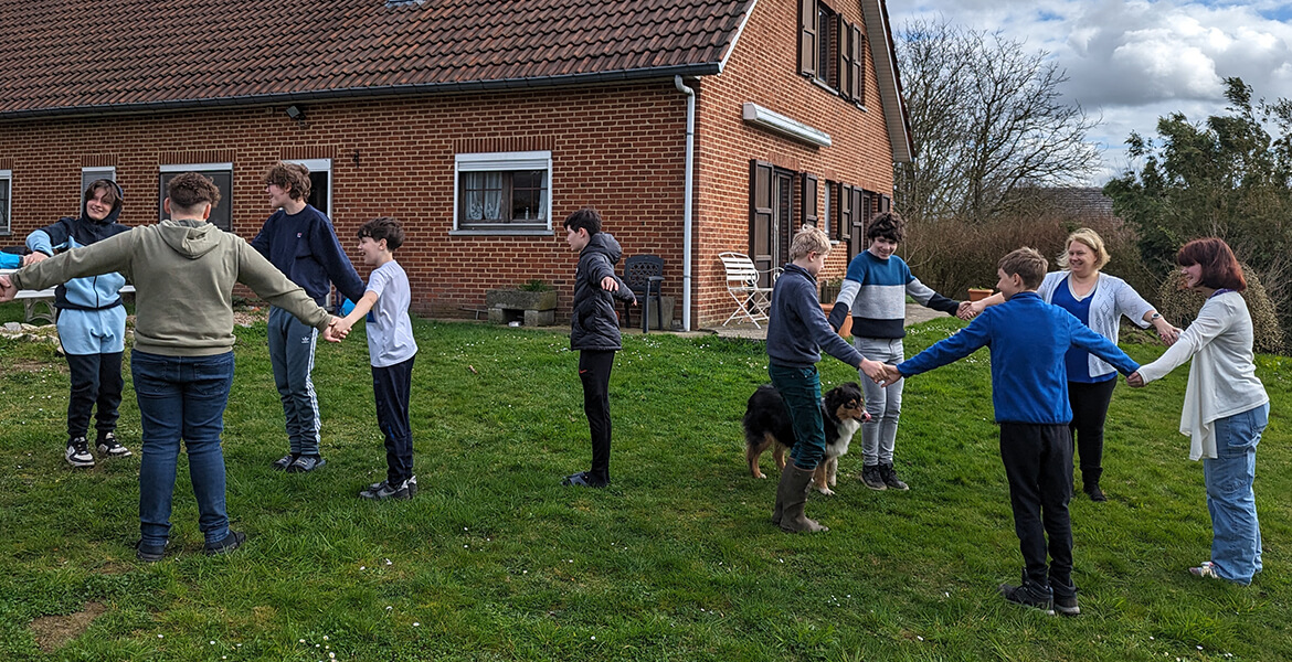 L'image montre un groupe d'élèves à l'extérieur, devant une maison en briques, participant à une activité pratique pour apprendre les mathématiques, plus précisément la symétrie orthogonale. Les élèves sont en cercle et semblent interagir les uns avec les autres, peut-être en utilisant des objets ou des repères au sol pour visualiser et comprendre les concepts de symétrie. Un chien est également présent au centre du cercle, ajoutant une touche de convivialité à la scène.